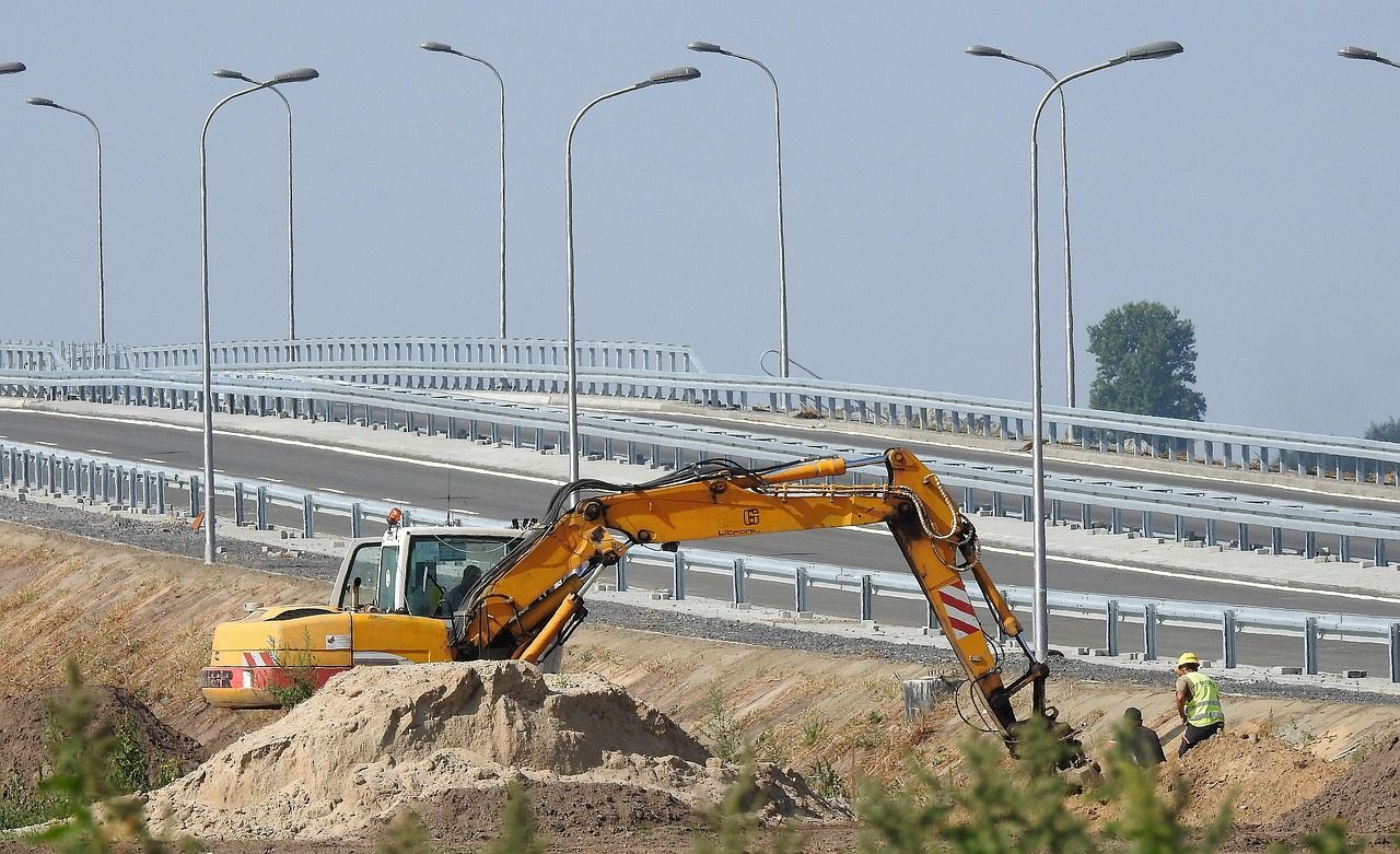 Koparka kopie wykop wzdłuż drogi, obok pracują robotnicy
